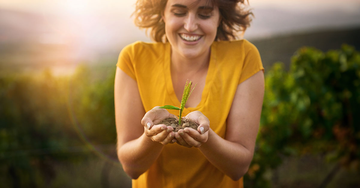 A person in a yellow shirt is smiling while holding a small plant with soil in their cupped hands, embodying eco-friendly design tips. The background features a blurred, sunny outdoor setting with lush greenery.